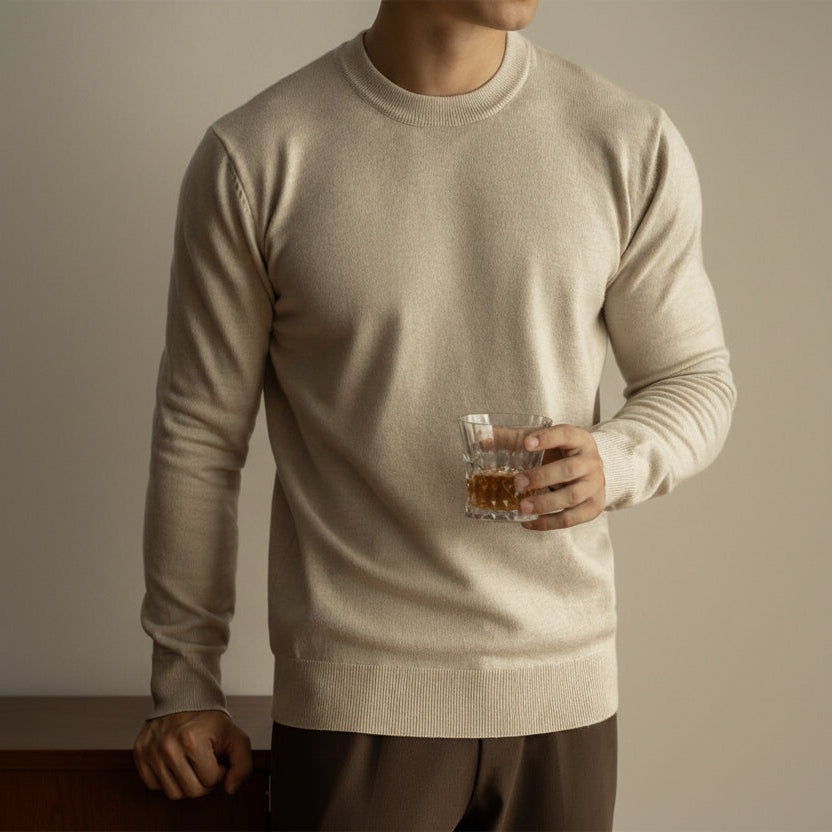 Man wearing a beige sweater holding a glass of whiskey against a neutral background